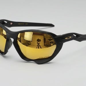 May include: Black sunglasses featuring a matte frame and reflective gold-toned lenses. The lenses are marked with the text PRIZM. The temples display a small gold-colored logo emblem. The interior of the temple includes the text USA and 128.