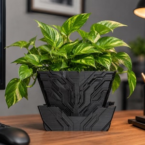 Pode incluir: Um vaso preto impresso em 3D com um padrão geométrico em relevo de inspiração tecnológica no exterior. O vaso contém uma exuberante planta pothos variegada com folhas verdes e amarelas em forma de coração que caem pelas bordas superiores.