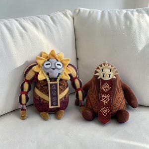 Puede incluir: Dos peluches con personajes estilizados. La figura de la izquierda tiene una cabeza amarilla en forma de sol, brazos a rayas y un colgante rectangular con símbolos. La figura de la derecha es marrón, redondeada, con cara solar y panel rojo.