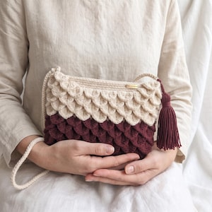 May include: A crochet crossbody bag featuring a textured scale pattern in cream and deep burgundy. The bag includes a matching cream-colored strap and a prominent deep burgundy tassel attached to the side. A small gold-toned rectangular tag is visible.