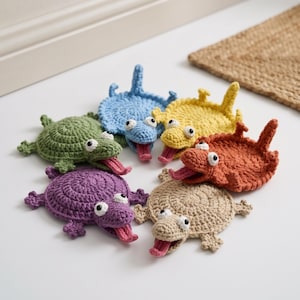 Könnte beinhalten: Eine Sammlung von sechs gehäkelten Untersetzern in Schildkrötenform in den Farben Grün, Blau, Gelb, Orange, Violett und Beige. Jede Schildkröte hat einen runden Panzer, vier kleine Beine, einen Schwanz, einen Kopf mit zwei weißen Augen und eine herausgestreckte rosa Zunge.