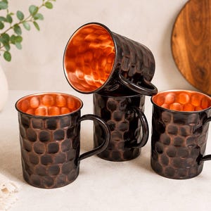 May include: A set of four mugs featuring a dark, hammered exterior finish and a bright, polished copper interior. Each mug has a sturdy handle attached to the side, with one mug positioned horizontally on top of the others in a stacked arrangement.