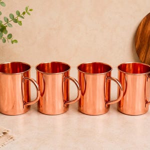 May include: A set of four polished copper mugs, each featuring a cylindrical body and a curved side handle. The mugs have a smooth, reflective metallic finish and are arranged in a horizontal row, showcasing their uniform design and warm colour.