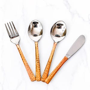 May include: A four-piece flatware set consisting of a fork, two spoons, and a butter knife. Each piece features a polished silver-tone metal head and a textured, hammered copper-colored handle that tapers toward the end of the utensil.