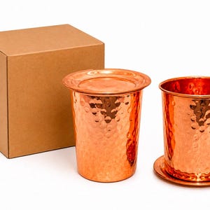 Peut inclure: Deux gobelets en cuivre martelé avec une finition métallique brillante. Un gobelet est recouvert d'un couvercle circulaire plat, tandis que l'autre repose sur un dessous de verre circulaire assorti. Une boîte en carton brun est placée derrière.