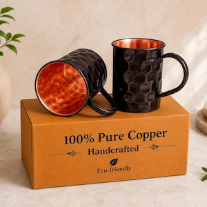 May include: Two black hammered mugs with metallic copper interiors and handles, resting on a brown cardboard box labelled 100% Pure Copper Handcrafted Eco-friendly. The mugs feature a textured exterior surface and a smooth, polished copper finish inside.