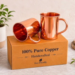 Peut inclure: Deux tasses en cuivre poli avec des anses incurvées posées sur une boîte en carton rectangulaire. La boîte porte l'inscription 100% Pure Copper, Handcrafted et Eco-friendly avec une icône de feuille. Les tasses ont une finition métallique lisse.