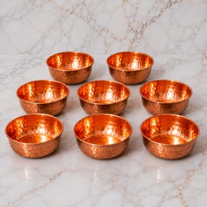 May include: A set of eight small, round bowls crafted from polished copper. Each bowl features a distinct hammered texture across its exterior surface, creating a reflective, metallic appearance with warm orange and brown tones throughout.