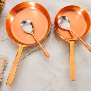 Puede incluir: Dos sartenes redondas de cobre con bordes martillados y mangos largos de metal dorado. Cada sartén contiene una cuchara de metal plateado pulido con mango texturizado. Las sartenes varían en tamaño, la más grande está a la izquierda.