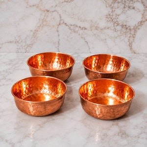 May include: A set of four round bowls crafted from polished copper, each featuring a distinct hammered texture across the exterior and interior surfaces. The bowls have a wide, open design with smooth, rounded rims and a warm, metallic orange finish.