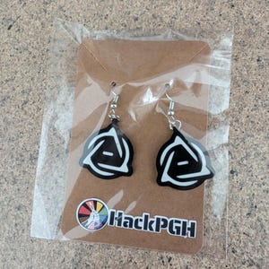 May include: A pair of dangle earrings featuring a black and white triangular logo design with silver-toned metal hooks. The earrings are attached to a brown cardboard display card that includes a circular logo and the text HackPGH in black.