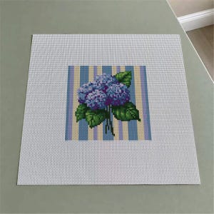 May include: A square needlepoint canvas featuring a central design of blue and purple hydrangea flowers with green leaves. The floral motif is set against a background of vertical stripes in shades of light blue, yellow, and white, all on a white grid.