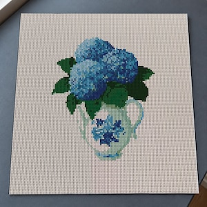 May include: A cross-stitch embroidery pattern on a square piece of light beige fabric. The design features a light blue pitcher with a dark blue floral motif, containing a cluster of three vibrant blue hydrangea blossoms with dark green leaves.