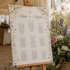 Può includere: Un tableau de mariage bianco su un cavalletto in legno, con i nomi Nina e Kevin, un monogramma e le liste degli ospiti per otto tavoli. Il pannello è decorato con un delicato bordo floreale ad acquerello di fiori selvatici viola e gialli.
