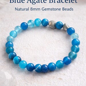 May include: A blue agate bracelet featuring 8mm gemstone beads in various shades of blue, ranging from light to deep navy. The bracelet includes two silver-toned metal accent beads with a textured, scale-like pattern, arranged in a circular loop.