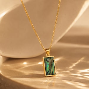 Puede incluir: Un collar de tono dorado con un colgante rectangular que presenta una incrustación de concha de abulón iridiscente. El colgante cuelga de una delicada cadena dorada, mostrando una vibrante paleta de colores verdes y azules en el patrón natural.