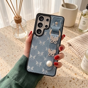 May include: A blue denim-textured smartphone case featuring a repeating white butterfly pattern. The case includes a matching blue denim strap attached with a gold-tone ring, adorned with two silver butterfly charms and a single white pearl bead.