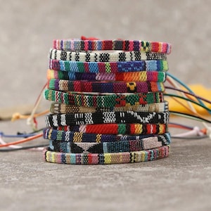 Peut inclure: Une pile verticale de onze bracelets en tissu tissé présentant divers motifs géométriques et rayures colorés. Chaque bracelet est fabriqué à partir de fils multicolores et comprend de fines ficelles lâches s'étendant sur les côtés de la pile.