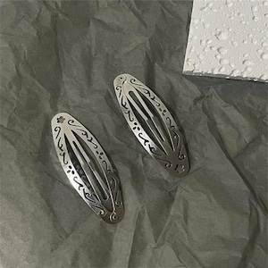 May include: Two oval-shaped silver-toned metal hair clips featuring intricate cut-out scrollwork patterns and small flower motifs. These hair accessories have a snap-style closure mechanism and a smooth, reflective finish on the surface.