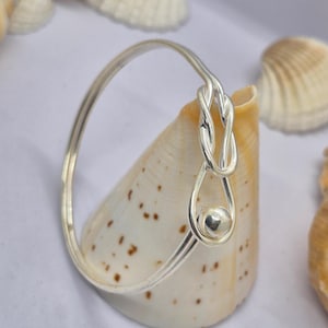 May include: A polished silver bangle bracelet featuring a continuous thin metal band that loops into a decorative knot design at one end, terminating in a small, smooth, spherical silver bead that rests within the lower curve of the knot.