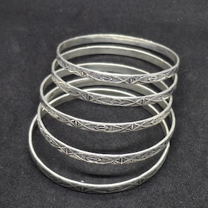 Puede incluir: Un conjunto de cinco brazaletes plateados apilados. Cada pulsera circular presenta un patrón geométrico grabado repetitivo que consiste en líneas que se cruzan y pequeñas formas de diamante a lo largo de la superficie exterior.