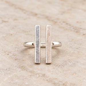 Puede incluir: Un anillo de tono plateado que presenta dos barras rectangulares verticales y paralelas que se extienden hacia arriba desde la banda. La superficie metálica tiene un acabado mate ligeramente texturizado, creando una apariencia limpia y geométrica. El anillo está construido a partir de una sola pieza de metal que forma la banda y las dos barras verticales.