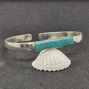 Op de afbeelding: Een zilveren armband met een rechthoekige, horizontaal georiënteerde turkoois steen met donkere, complexe aders. De metalen band is glad en gepolijst, met een open achterkant, wat zorgt voor een strakke, metalen afwerking en een levendige blauwgroene steen.