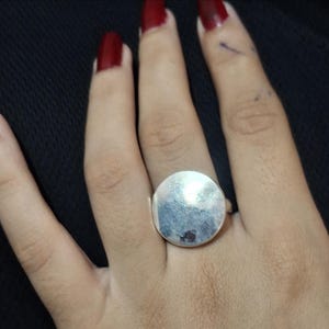 Op de afbeelding: Een zilverkleurige ring met een grote, platte, cirkelvormige schijf als kop. Het oppervlak van de schijf is glad en reflecterend en vangt het licht op over de ronde vorm. De ringband is dun en verbonden met de achterkant van het middelpunt.