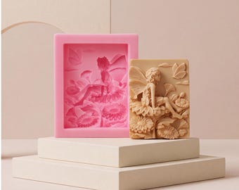 Pode incluir: Um molde de silicone rosa retangular com um design rebaixado de uma fada sentada sobre uma flor, acompanhado por uma barra de sabão retangular bege moldada, exibindo um relevo da fada, flores e uma borboleta em sua superfície.