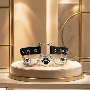 May include: A black leather-like choker featuring silver-toned hardware, including a central cat-ear shaped ring, a dangling black and silver paw print charm, and two small silver bells suspended by chains from the band with circular studs.
