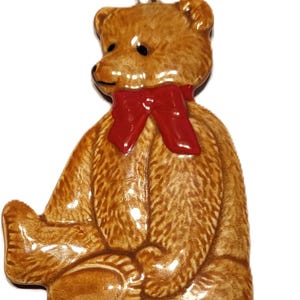 May include: A ceramic teddy bear ornament with a red bow tie. The bear is sitting with its legs extended and has a gold string for hanging. The bear is brown with black eyes and a glossy finish.