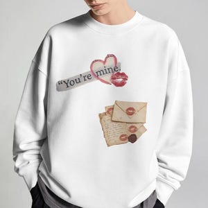 May include: A white long-sleeve sweatshirt featuring a graphic print of a red heart, a red lipstick kiss mark, and the text "You're mine." Below this, a stack of vintage-style envelopes and letters decorated with red lipstick kiss marks.