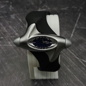 May include: A wristwatch featuring a dark blue oval dial encased in a matte silver-toned metal frame with pointed side accents. The watch is attached to a flexible black rubber strap that wraps around a white cylindrical display stand.
