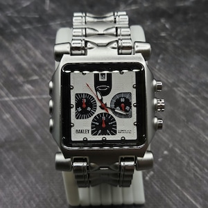 May include: A silver-toned Oakley wristwatch with a rectangular face, featuring a white dial with three black sub-dials, red hands, and a date window at the top. The watch has a metallic link bracelet and includes the text Oakley and Swiss Made.