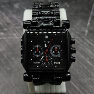 May include: A black square-faced wristwatch with a matching black metal link band. The watch features a black dial with three sub-dials, red hands, a date window at the top, and the Oakley logo printed in white on the lower left side of the face.