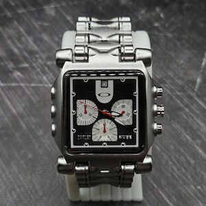 May include: A silver-tone stainless steel wristwatch with a rectangular black dial featuring three white sub-dials, red hands, and the Oakley logo. The watch includes a date window at the twelve o'clock position and a metallic link bracelet band.