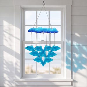 May include: A hanging mobile featuring a horizontal blue glass wave-shaped top piece suspended by thin wires. Below the wave, eight translucent light blue glass dolphin figures are arranged in a tiered formation, hanging from the top piece.