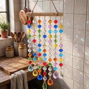 Puede incluir: Un atrapasol colgante que presenta siete hilos verticales de cuentas de cristal facetado multicolor suspendidos de un listón de madera horizontal. Las cuentas están dispuestas en varios colores, incluyendo rojo, azul, amarillo, verde y rosa, con colgantes de cristal en forma de gota y esfera en la parte inferior.