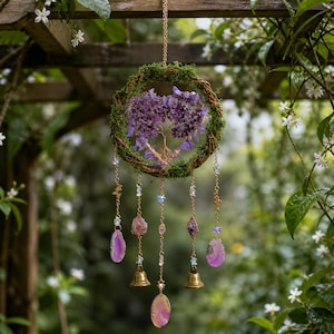 Può includere: Una decorazione da parete circolare composta da una corona di vite ricoperta di muschio con un albero centrale in filo metallico e frammenti di ametista viola. Cinque catenelle dorate pendono dal basso, decorate con cristalli e due campanelli.