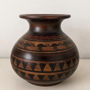 Peut inclure: Un vase rond en bois brun foncé avec un col large et un bord évasé. La surface présente des motifs géométriques peints à la main en noir et rouge, incluant une bande de silhouettes d'oiseaux stylisées et des rangées de triangles noirs.