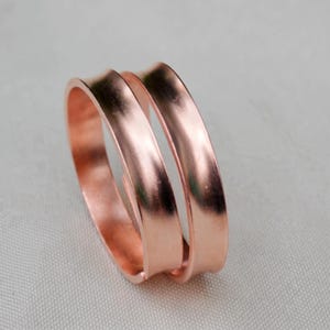 May include: A polished copper ring featuring a double-band design with a smooth, reflective surface. The ring has a continuous, open-ended structure that creates two parallel bands, showcasing the warm, metallic reddish-brown hue of the copper.