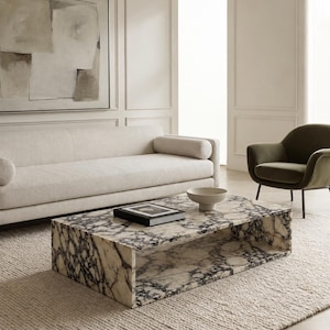 May include: A rectangular coffee table crafted from white marble with prominent dark grey veining. The table features a hollow, open-shelf design. A small, shallow off-white bowl and a black-covered book are placed on the flat top surface.