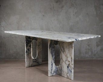 May include: A rectangular dining table crafted from white marble with prominent grey and dark blue veining. The table features a flat top supported by two vertical slab legs, each containing a central elongated oval cutout opening.