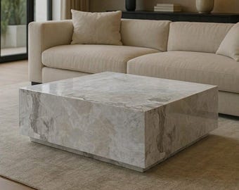 May include: A square coffee table featuring a polished light grey and white marble surface with natural veining patterns. The table has a low-profile, block-like design with a recessed base, creating a floating appearance. The stone surface is smooth.