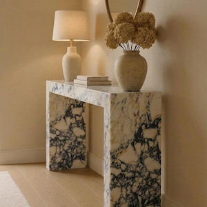 Peut inclure: Une console rectangulaire en marbre blanc avec des veines noires marquées. Sur la table se trouvent une lampe à poser en céramique blanche avec un abat-jour en tissu, deux livres empilés et un vase beige texturé rempli d'hortensias séchés.
