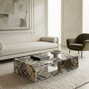 May include: A rectangular coffee table crafted from white marble with prominent dark grey and black veining. The table features a hollow centre section and a smooth, polished surface, topped with a single book and a small, shallow ceramic bowl.