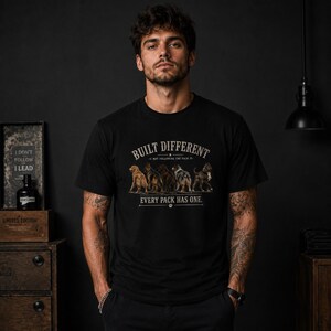May include: A black short-sleeved t-shirt featuring a graphic of five different dog breeds standing in a row. The text reads BUILT DIFFERENT, NOT FOLLOWING THE PACK, and EVERY PACK HAS ONE, with a small paw print icon centered at the bottom.