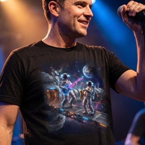 May include: A black t-shirt featuring a graphic print of two astronauts on a space station walkway. One astronaut plays a glowing blue and pink electric guitar, while the other plays a gold saxophone. The background shows deep space and Earth.