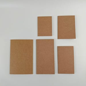 May include: A collection of five rectangular notebooks with plain, light brown covers. The notebooks are arranged in a group, featuring varying sizes and proportions, all displaying a simple, uniform, and unadorned paper texture.