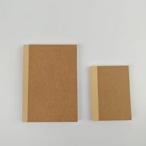 May include: Two rectangular notebooks with brown covers and a light beige vertical spine strip. The notebooks are positioned side by side, with one being significantly larger than the other, both featuring a plain, unadorned paper surface.
